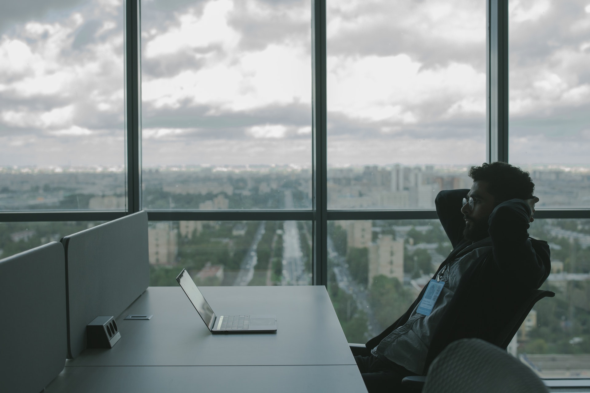 A man at a white desk leans back in his chair while staring at his laptop. A large window is behind him.