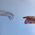 A robot hand and a human hand reach for each other against a light blue background.