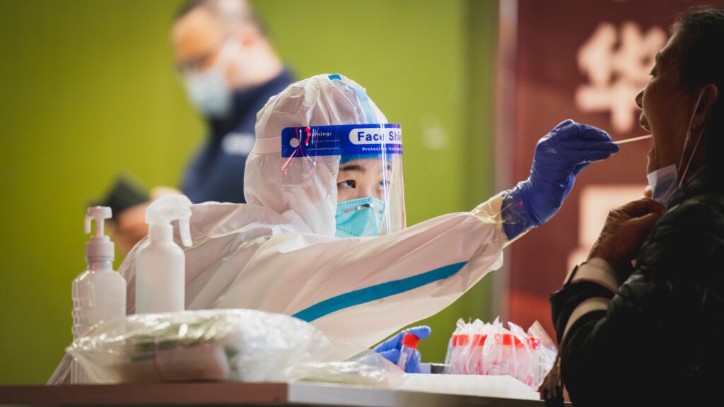 A healthcare worker in a biohazard suit, mask, and face shield tests an unseen patient for coronavirus with a mouth swab. Another helathcare worker in a mask stands behind her, against a green wall.
