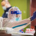 A healthcare worker in a biohazard suit, mask, and face shield tests an unseen patient for coronavirus with a mouth swab. Another helathcare worker in a mask stands behind her, against a green wall.