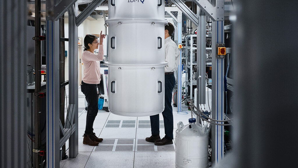 A large, cylindrical white container hangs from a metal frame in lab. A man is half-hidden behind it, while a woman in black pants and a pink shirt points to it.