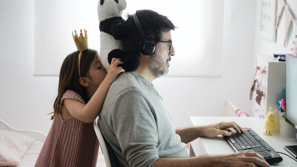 A father is working at home on a laptop at a desk. His daughter, wearing a pink dress and a crown, balances her stuffed panda on his shoulders while he tries to work.