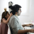 A father is working at home on a laptop at a desk. His daughter, wearing a pink dress and a crown, balances her stuffed panda on his shoulders while he tries to work.