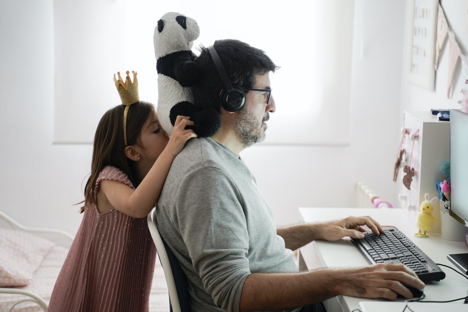 A father is working at home on a laptop at a desk. His daughter, wearing a pink dress and a crown, balances her stuffed panda on his shoulders while he tries to work.