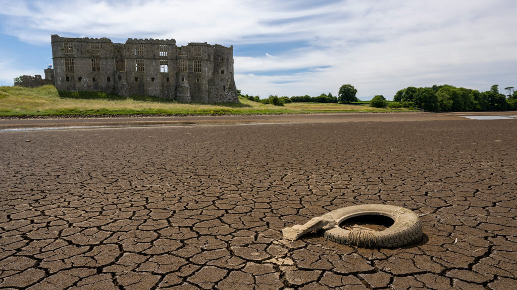 An old, dirty, deflated tire lies on a cracked river bed with no water. A stone castle is in the background on a green hill.