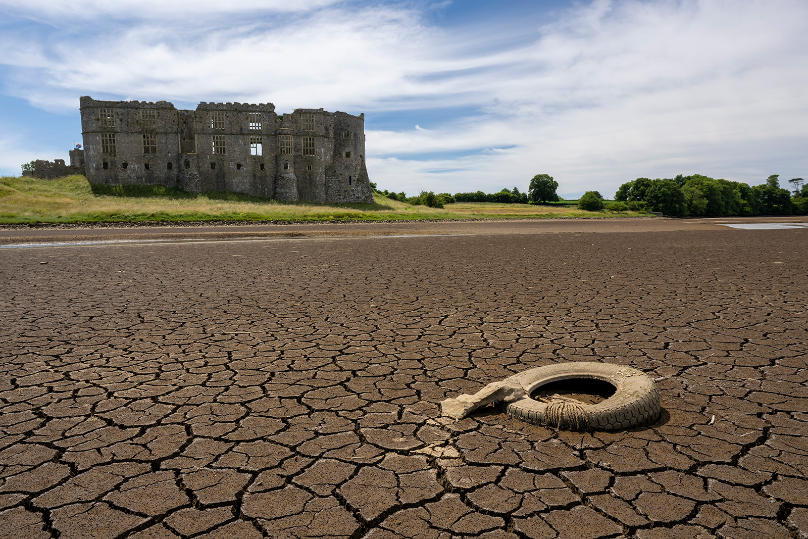 An old, dirty, deflated tire lies on a cracked river bed with no water. A stone castle is in the background on a green hill.
