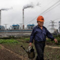 A man in a blue shirt with an orange hard hat is holding a water can as he walks across a field. Behind him are smoking chimneys from a factory.