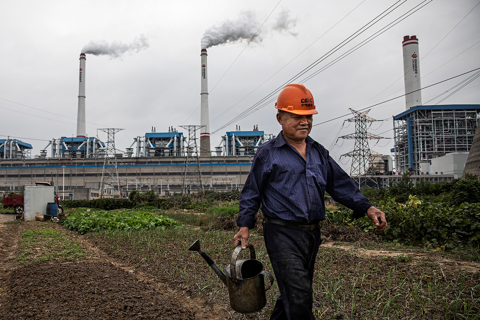 A man in a blue shirt with an orange hard hat is holding a water can as he walks across a field. Behind him are smoking chimneys from a factory.