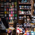 A man sits in front of a colorful stall, with shoes and spools of thread for sale. He holds his head in his hands.