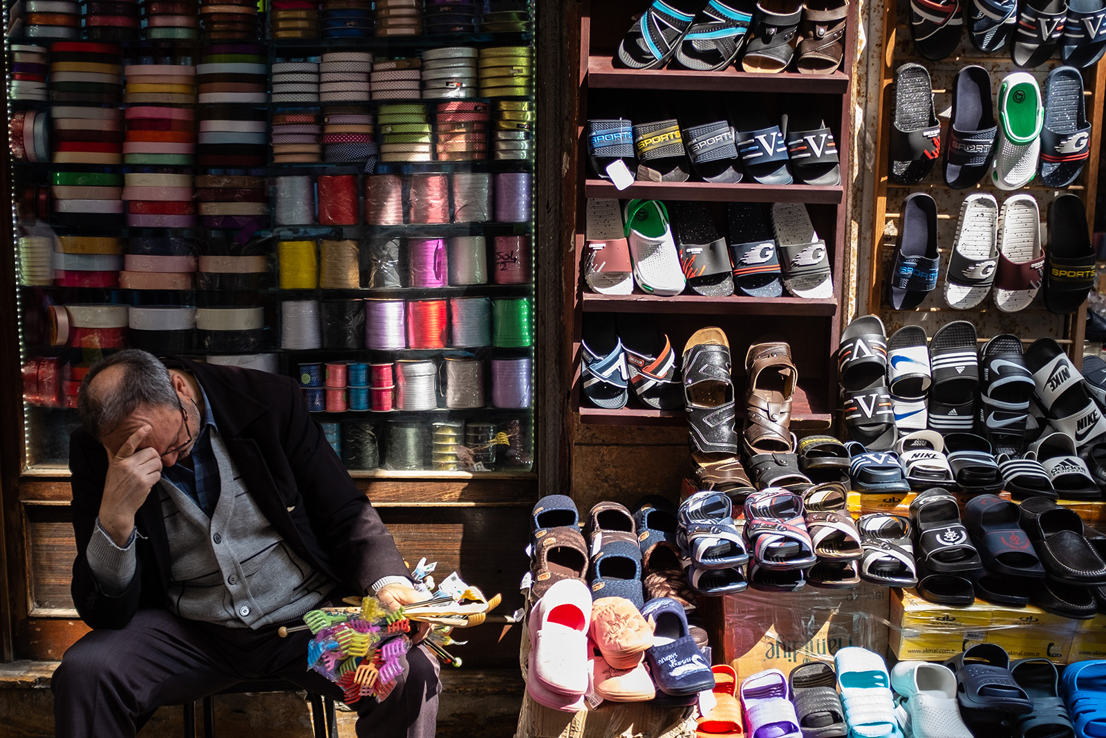 A man sits in front of a colorful stall, with shoes and spools of thread for sale. He holds his head in his hands.