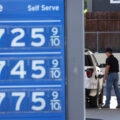 A blue display board with gas prices over $7. A man filling up a white car with gas can be seen in the background.