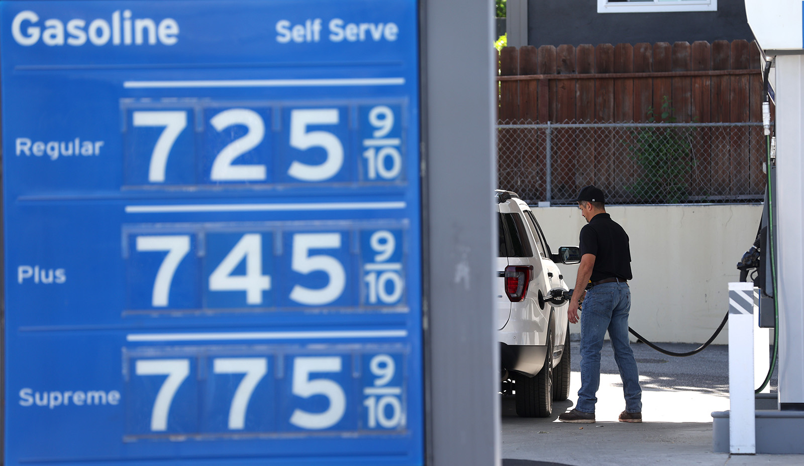 A blue display board with gas prices over $7. A man filling up a white car with gas can be seen in the background.
