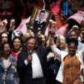 A man wearing a suit and glasses holds hands with a woman in a colorful dress and white cardigan. They both raise their held hands in triumph, as a crowd behind them cheers.