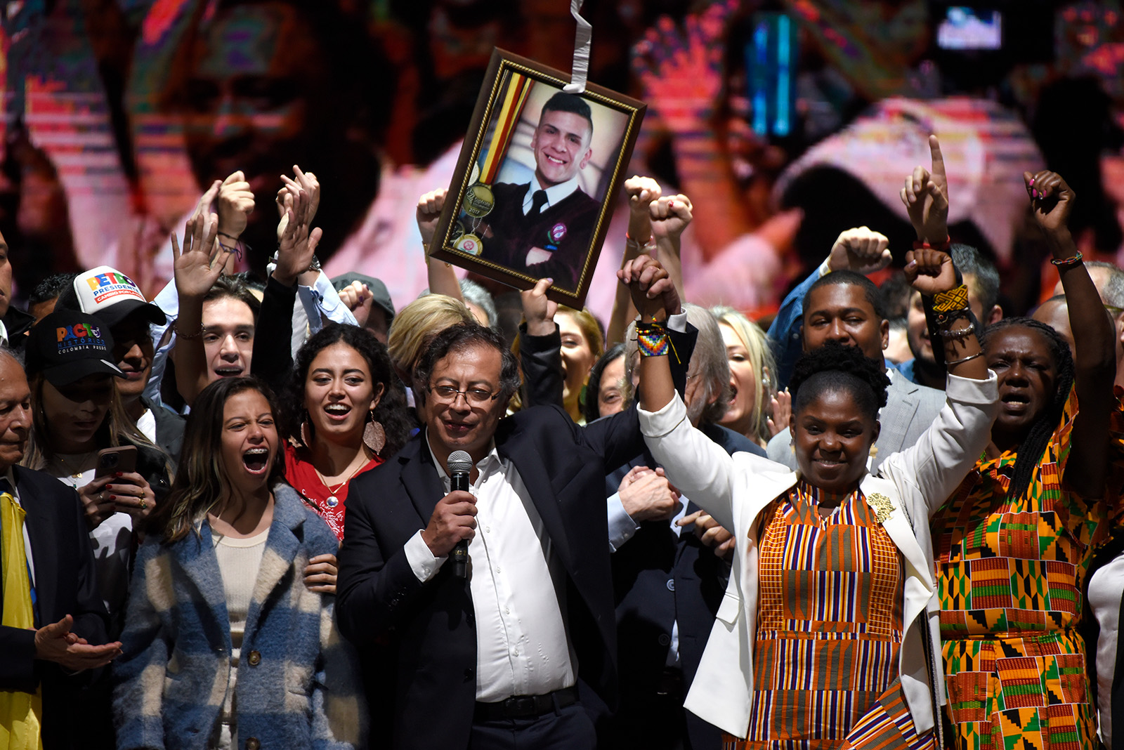 A man wearing a suit and glasses holds hands with a woman in a colorful dress and white cardigan. They both raise their held hands in triumph, as a crowd behind them cheers.
