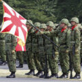 A group of soldiers in camouflage and wearing helmets stand in a group. A soldier in the foreground holds the Japanese flag.