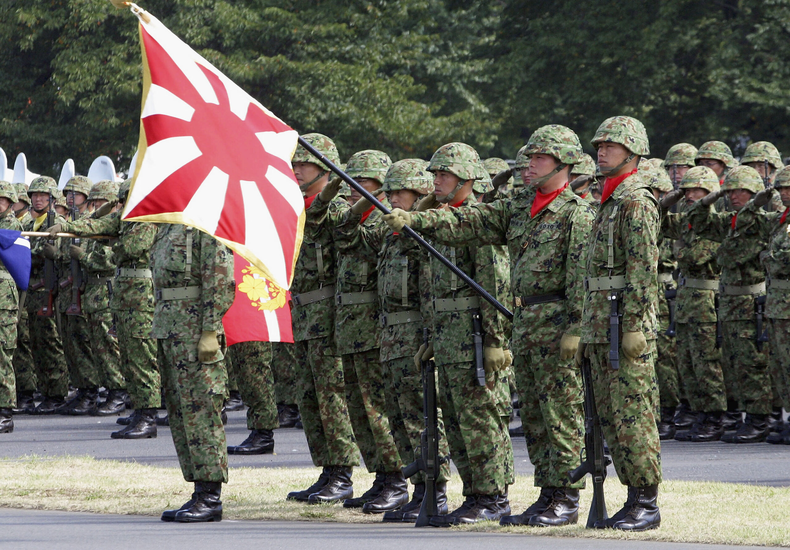 A group of soldiers in camouflage and wearing helmets stand in a group. A soldier in the foreground holds the Japanese flag.