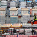 A woman in a yellow safety vest sits on an orange scooter-like vehicle as she moves through warehouse aisles of cartons of packaged food.