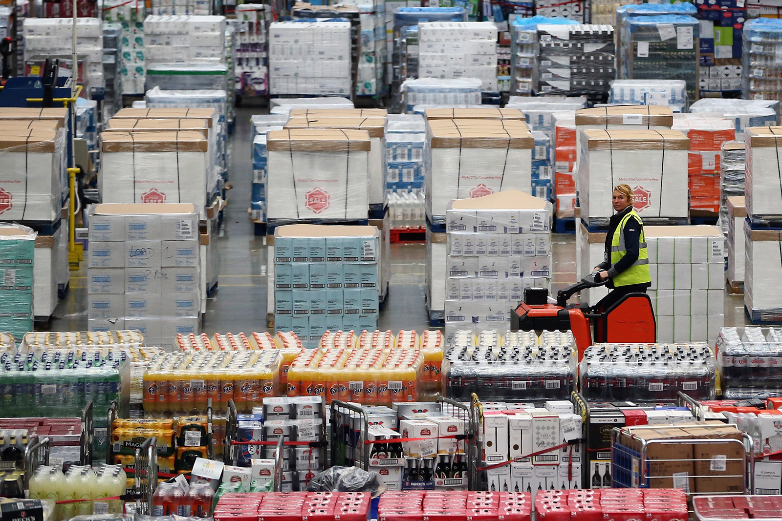 A woman in a yellow safety vest sits on an orange scooter-like vehicle as she moves through warehouse aisles of cartons of packaged food.