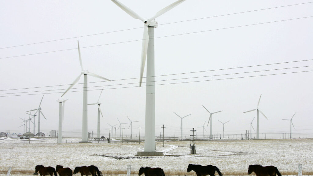 Wind turbines in a snowy field with an overcast grey sky. A line of brown ponies walks past the turbines in the foreground.