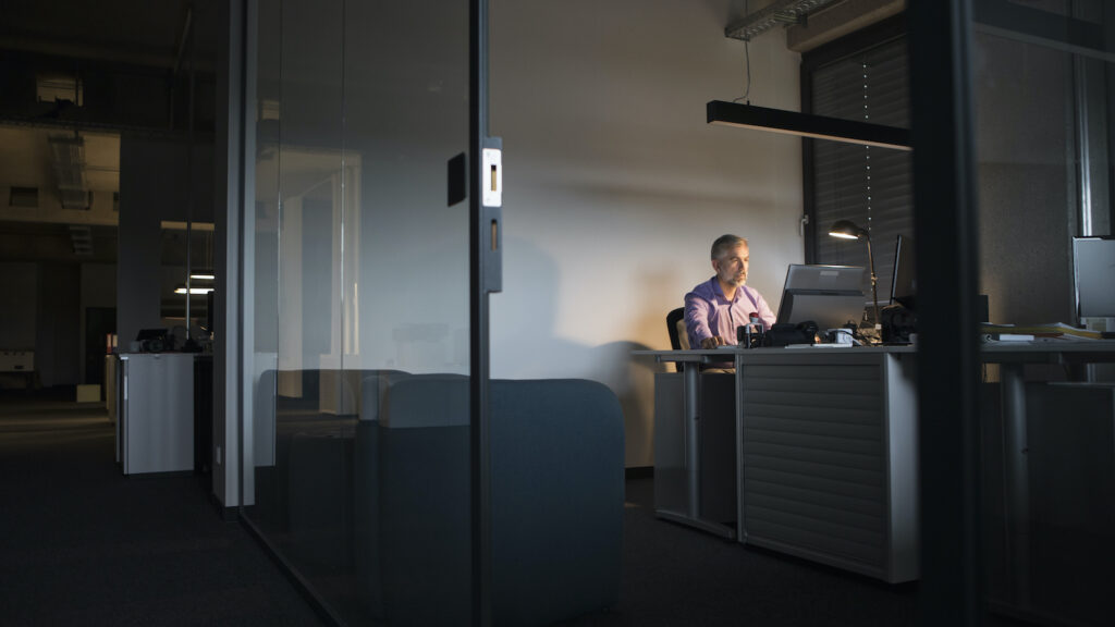 A business man sits in a dark office alone, with one light on above him.