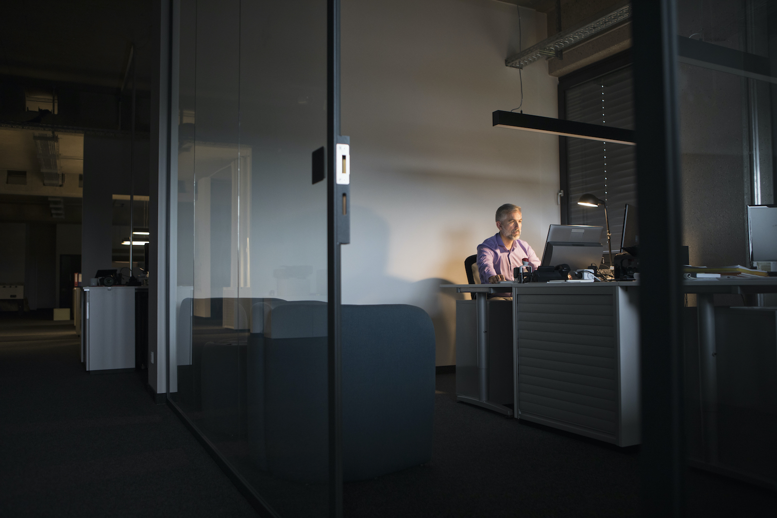 A business man sits in a dark office alone, with one light on above him.