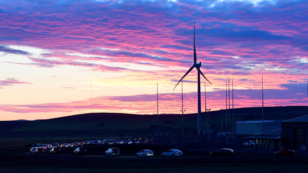 A wind turbine stands against a pink and purple sunrise. A line of cars can be seen below.