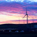 A wind turbine stands against a pink and purple sunrise. A line of cars can be seen below.