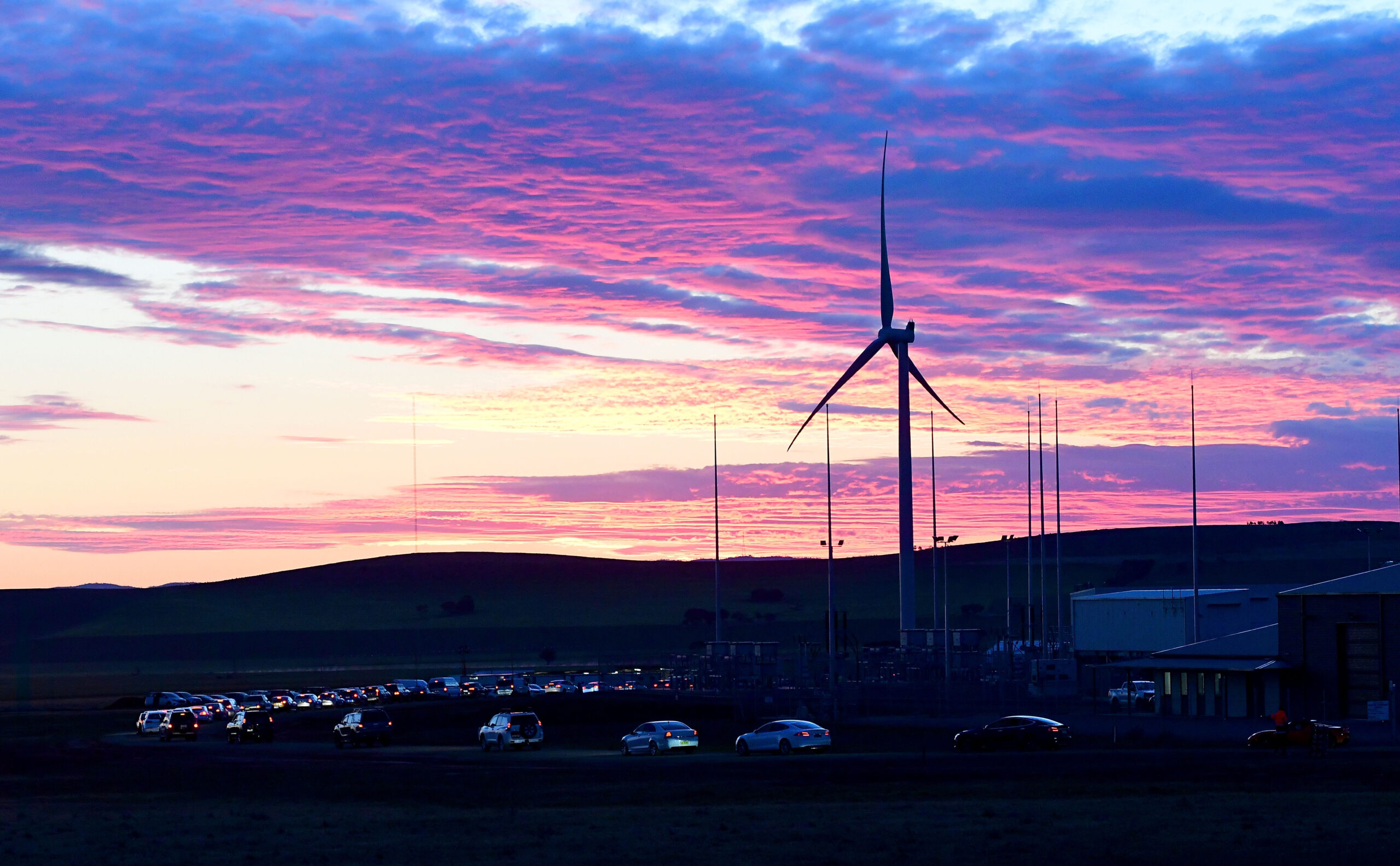 A wind turbine stands against a pink and purple sunrise. A line of cars can be seen below.