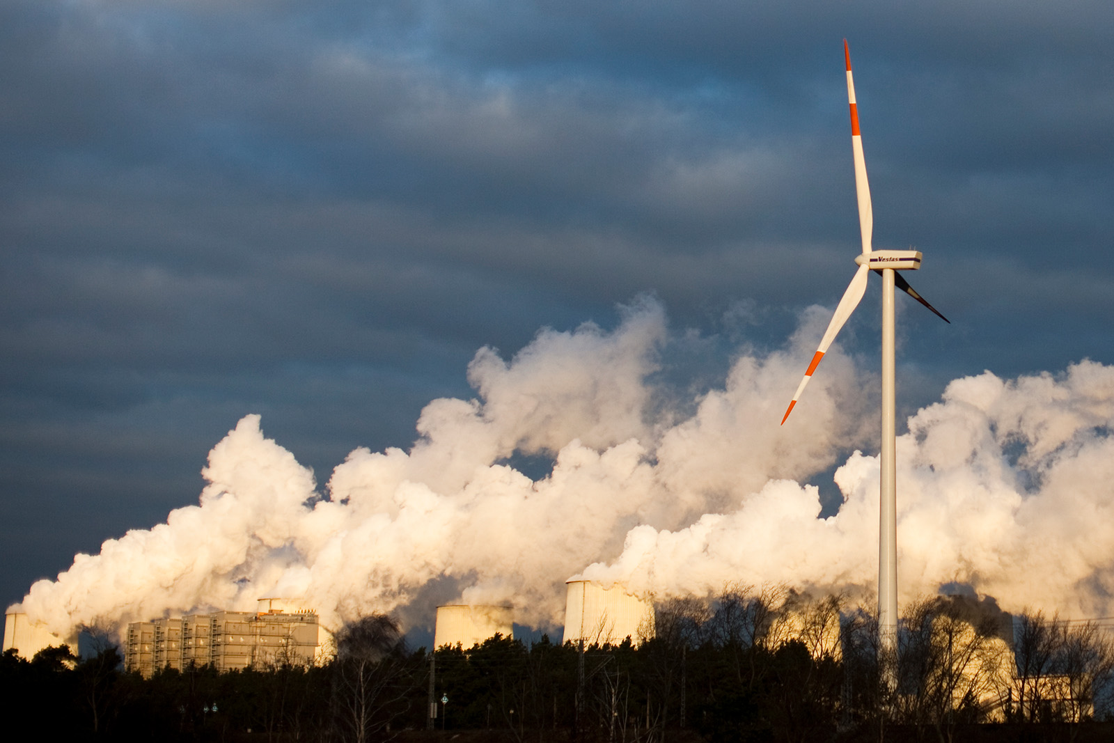 A wind turbine is in the foreground, on an overcast day. Behind it are plumes of white smoke from the chimneys of a power station, set among dark trees.