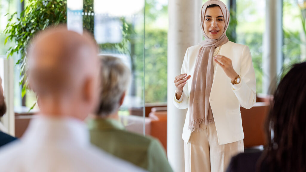 A woman wearing a hijab and a white pantsuit stands in front of a group and gives a presentation. There are windows behind her, through which can be seen trees.