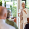 A woman wearing a hijab and a white pantsuit stands in front of a group and gives a presentation. There are windows behind her, through which can be seen trees.