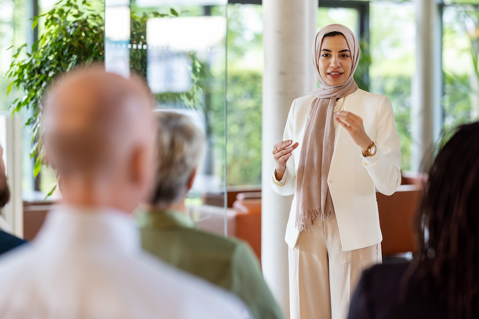 A woman wearing a hijab and a white pantsuit stands in front of a group and gives a presentation. There are windows behind her, through which can be seen trees.