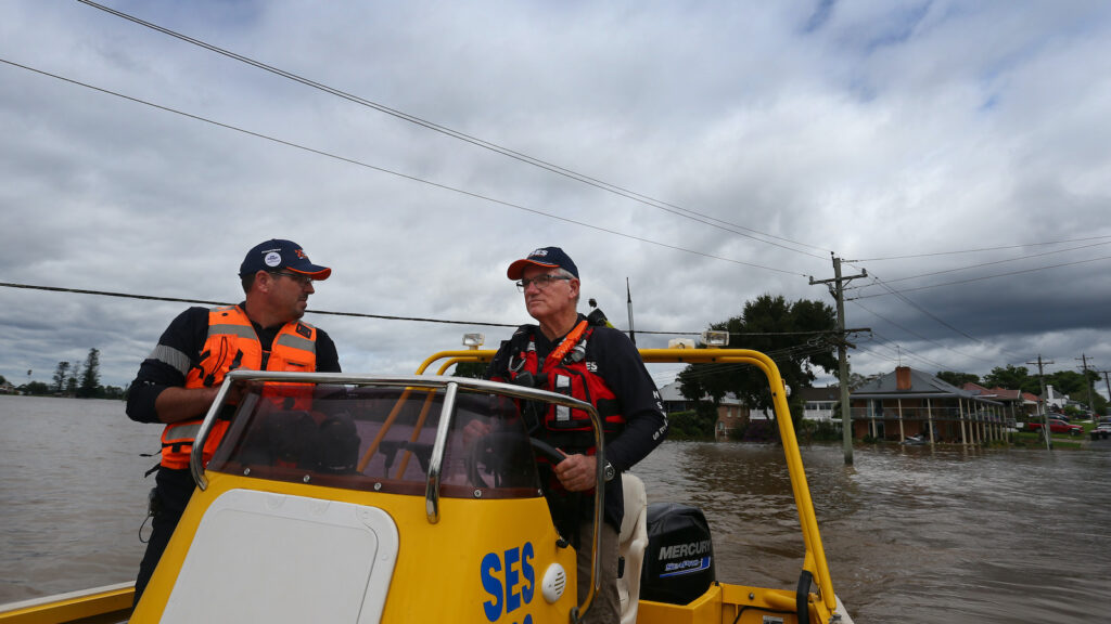 Two men wearing orange floatation vests and baseball caps ride in a yellow rescue boat down a flooded street. Houses nearly underwater can be seen in the background.