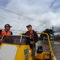 Two men wearing orange floatation vests and baseball caps ride in a yellow rescue boat down a flooded street. Houses nearly underwater can be seen in the background.