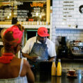 A barista behind a counter makes coffee, while a woman waits at the cash register for it.