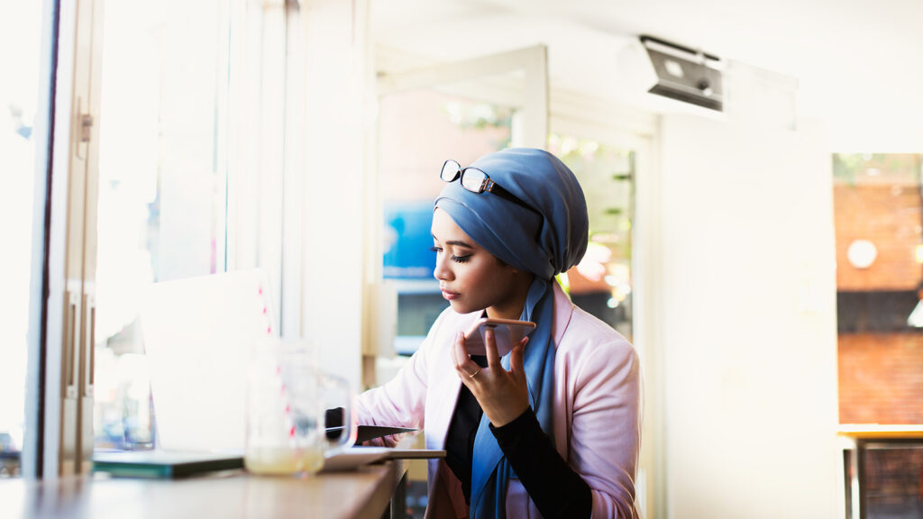 A young Muslim woman in a headwrap listens to her phone as she takes notes. She is sitting in a cafe.