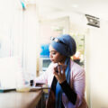 A young Muslim woman in a headwrap listens to her phone as she takes notes. She is sitting in a cafe.