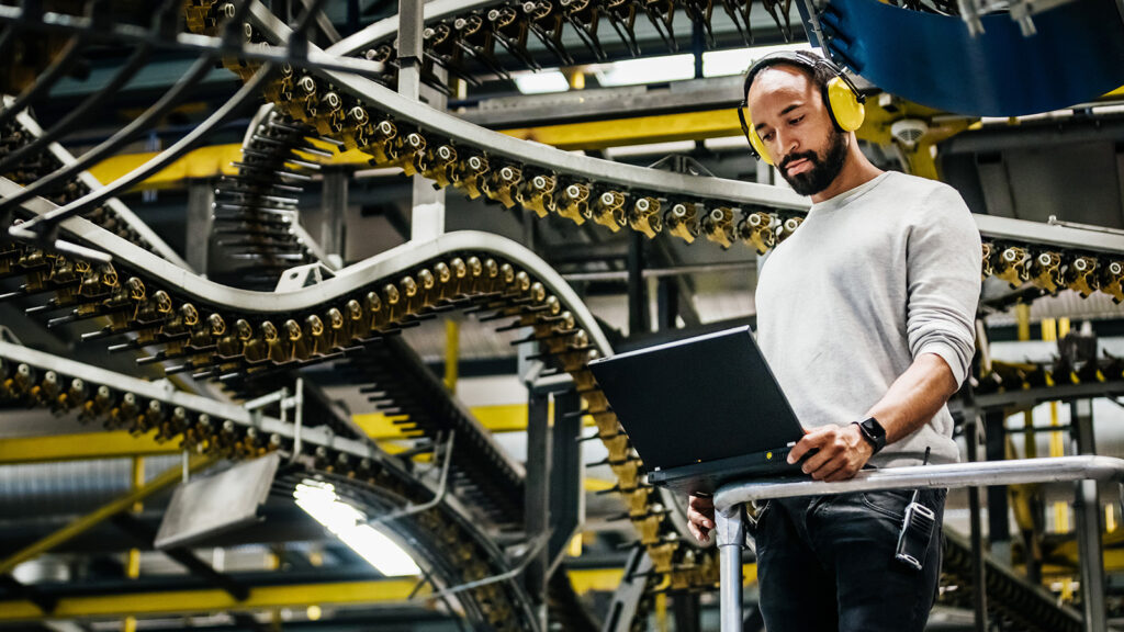 A bearded man wearing headphones stands working on a laptop computer while behind him is a series of conveyor belt loops.