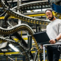 A bearded man wearing headphones stands working on a laptop computer while behind him is a series of conveyor belt loops.