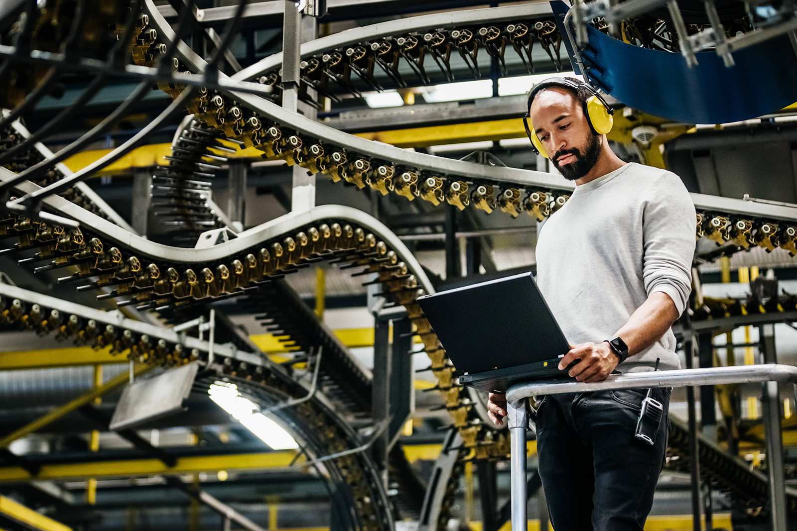 A bearded man wearing headphones stands working on a laptop computer while behind him is a series of conveyor belt loops.