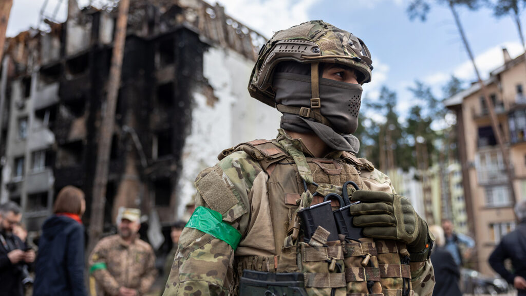 A soldier in camo and a mask stands in the foreground, a green armband on his arm. Behind him stands a destroyed building.