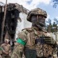 A soldier in camo and a mask stands in the foreground, a green armband on his arm. Behind him stands a destroyed building.