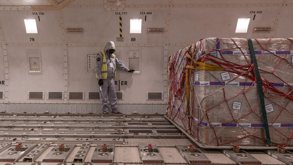 A worker in a white suit with a hood and yellow safety vest stares at their feet as they lean against a wall in a warehouse. Next to them is a large pallet of boxes wrapped in plastic, ready for shipping.
