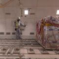 A worker in a white suit with a hood and yellow safety vest stares at their feet as they lean against a wall in a warehouse. Next to them is a large pallet of boxes wrapped in plastic, ready for shipping.