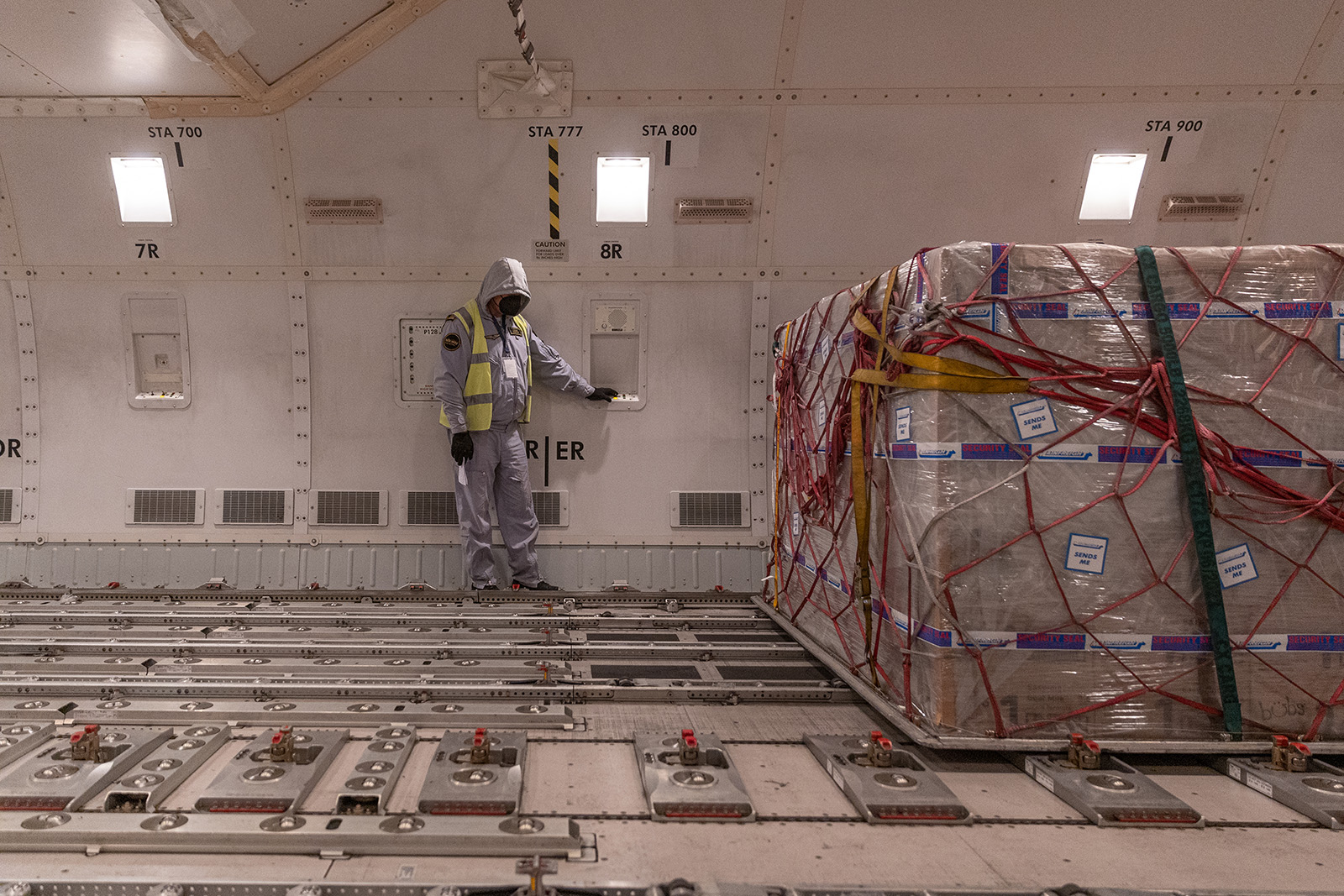 A worker in a white suit with a hood and yellow safety vest stares at their feet as they lean against a wall in a warehouse. Next to them is a large pallet of boxes wrapped in plastic, ready for shipping.