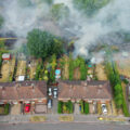 A birds-eye view of houses with green backyards. Smoke from wildfires rises from the fields behind them.