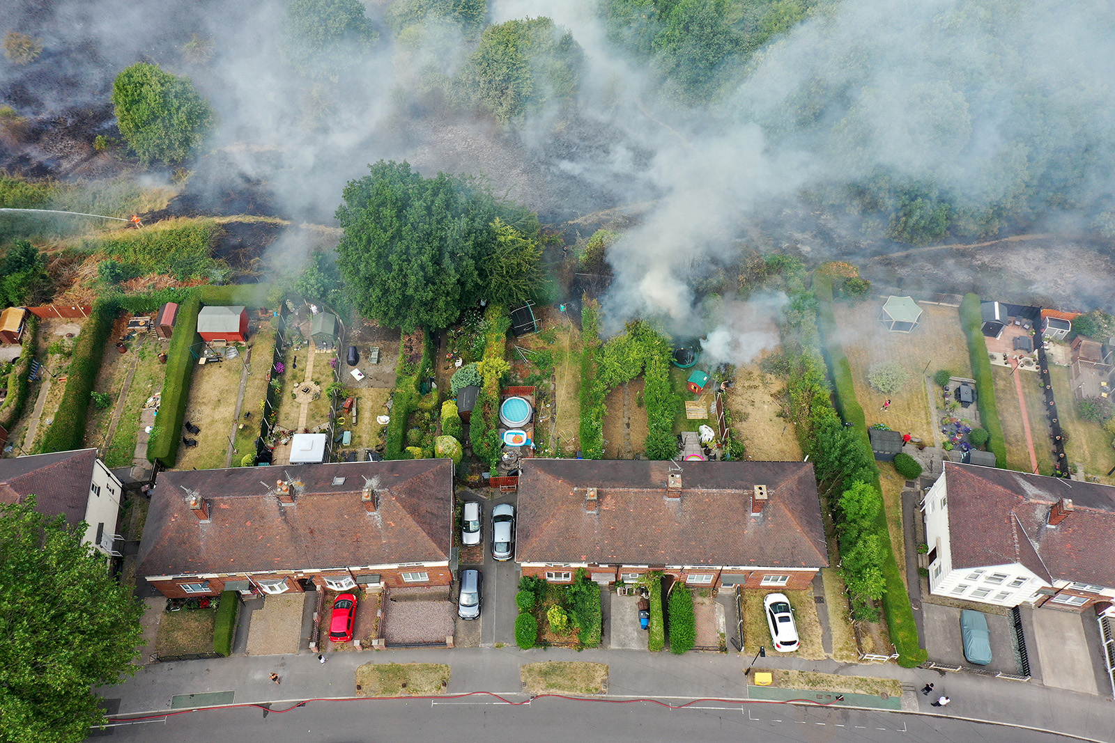 A birds-eye view of houses with green backyards. Smoke from wildfires rises from the fields behind them.