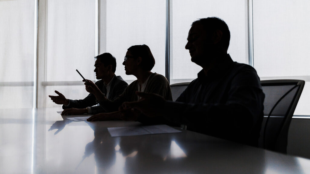Three shadowy silhouettes of people in profile sitting at an office table, with windows behind them.