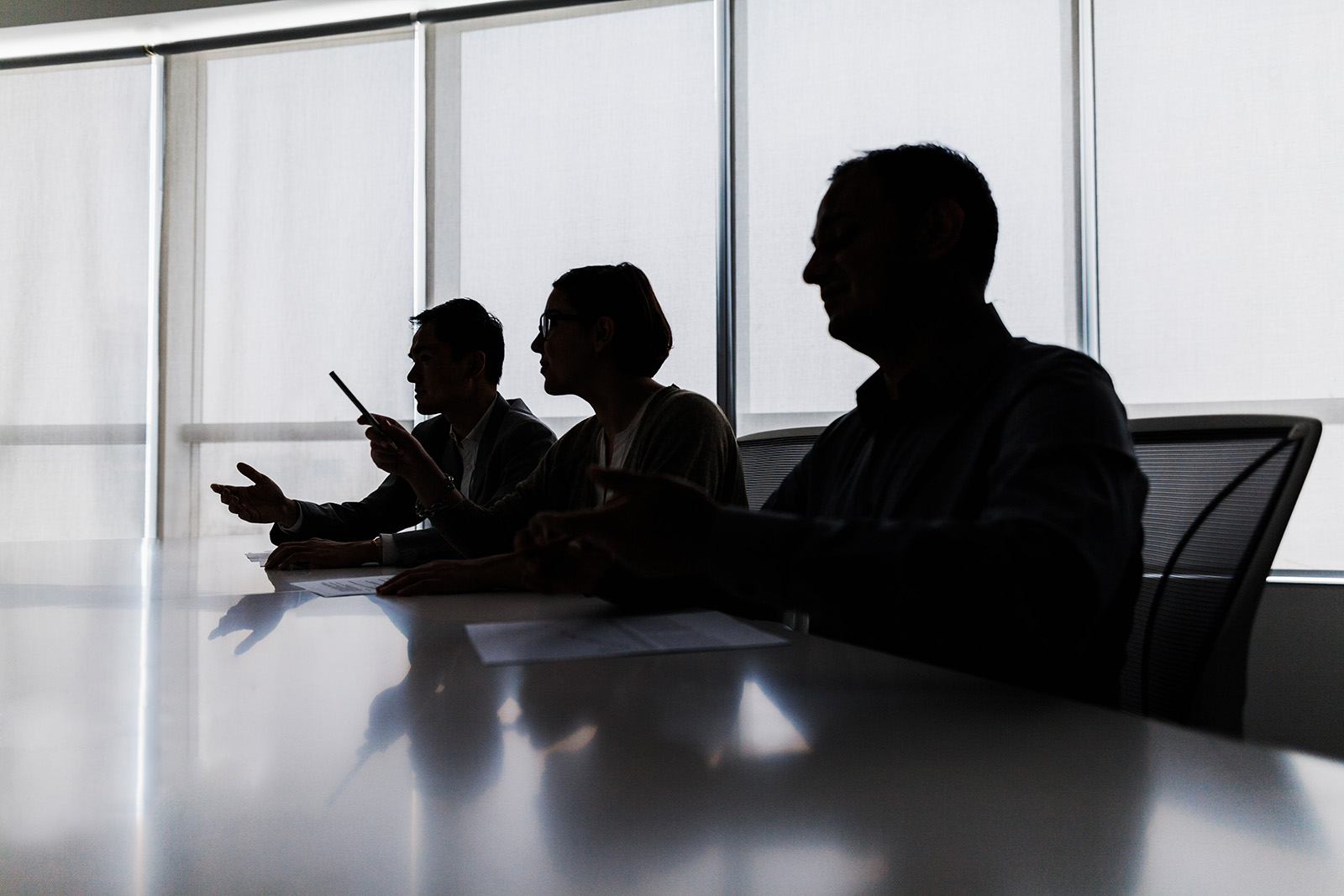 Three shadowy silhouettes of people in profile sitting at an office table, with windows behind them.