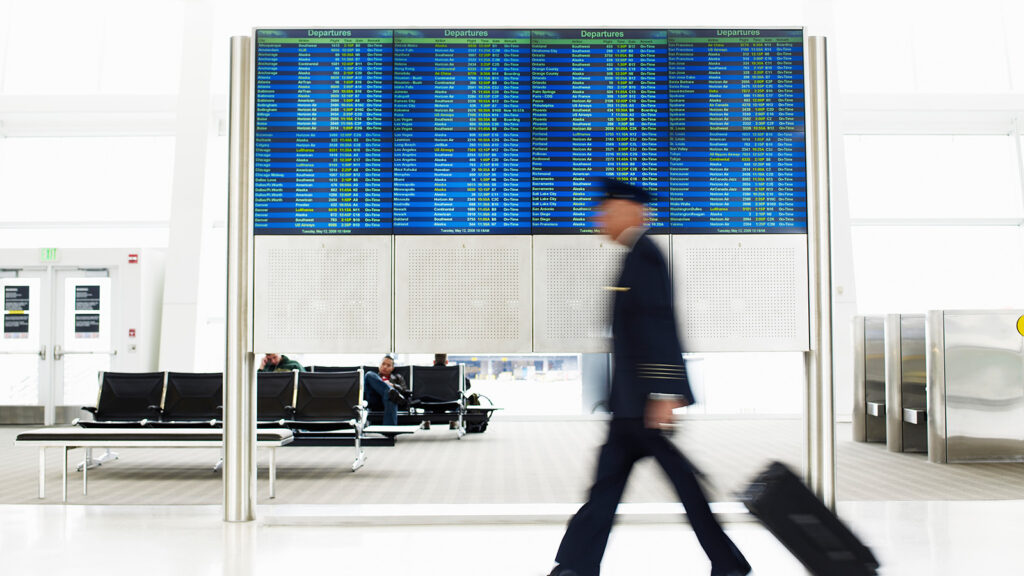 A blurred pilot walks past a board showing departures as he rolls a suitcase.